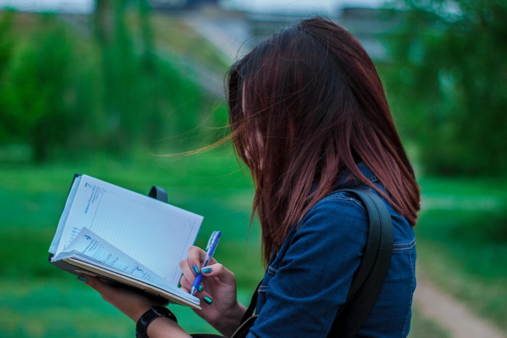 Side view of a young woman writing in a notebook while outdoors, capturing a thoughtful moment.